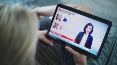 ZAPORIZHYA, UKRAINE - 7 Haziran 2024: Kanepede yatarken tablet bilgisayar kullanarak ABD seçimlerinin oylama sonuçlarını izleyen Kafkasyalı kadının görüntüsü. Biden ile anket sonuçları