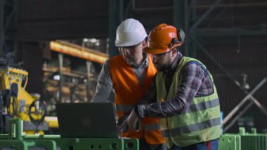 Üniformalı ve kasklı iki ağır endüstri mühendisi imalat fabrikasında dizüstü bilgisayar ve cep telefonu kullanarak çalışıyorlar. Olgun yönetici ve yetişkin çalışan fabrikada tartışıyorlar. Yavaş çekim.