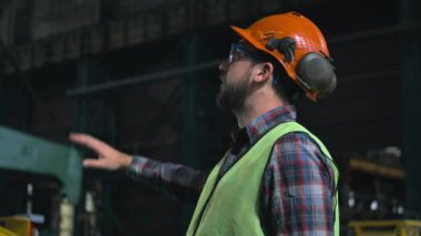 Erkek çalışan endüstriyel ekipman işletimini yönetir ve kontrol eder. Üretim fabrikasında koruyucu üniformalı ve miğferli profesyonel ağır sanayi teknisyeni veya mühendisi. Yavaş çekim.