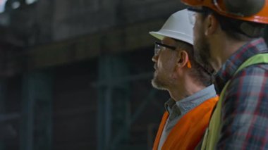 Güvenlik üniformalı iki profesyonel ağır endüstri teknisyeni ve iş sürecini tartışan ve imalat fabrikasındaki makine endüstrisi ekipmanlarına bakan kalın şapkalılar. Yavaş çekim.