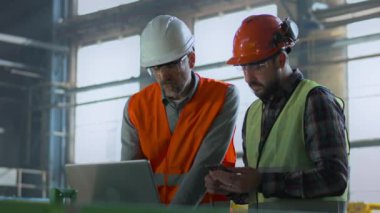 Güvenlik üniformalı ve kasklı iki endüstri teknisyeni depoda dizüstü bilgisayar ve cep telefonu üzerinde çalışıyorlar. Olgun mühendis ve yetişkin işçiler imalat fabrikasında konuşuyor. Yavaş çekim.