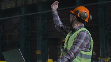 Üretim tesisi ya da fabrikada güvenlik üniforması giyen profesyonel bir ağır sanayi mühendisi. Erkek işçi endüstriyel ekipman işini yönetir ve dizüstü bilgisayar kullanır. Yavaş çekim.