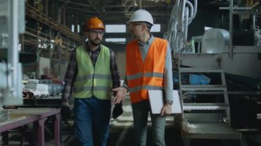 Güvenlik üniformalı iki ağır endüstri mühendisi ve sert şapkalı iki kişi üretim tesisinde yürüme sürecini tartışıyor. Olgun yönetici dizüstü bilgisayarı tutuyor, fabrikadaki yetişkin işçilerle konuşuyor. Yavaş çekim.