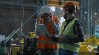 Üretim fabrikasında koruyucu üniforma ve kask giyen iki profesyonel endüstri teknisyeni duruyor. Olgun mühendis VR kulaklık kullanıyor. Yetişkin çalışan bilgisayarda çalışıyor. Yavaş çekim.