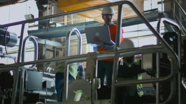 Profesyonel ağır endüstri, güvenlik üniforması, kask ve gözlüklü olgun mühendis dizüstü bilgisayar kullanır ve imalat fabrikasındaki teknisyenlerle konuşur. İki erkek çalışan ekipmanla çalışıyor. Yavaş çekim.