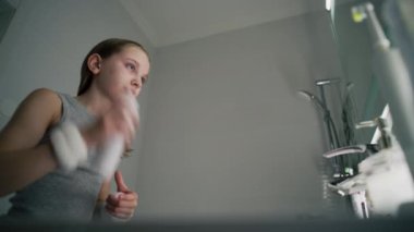 Genç kız banyodaki aynaya bakar ve yüzüne termal su püskürtür. Yumuşak yüzlü güzel kız lavabonun önünde duruyor. Sağlıklı cilt bakımı. Sabah banyosu konsepti.