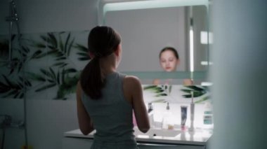 Genç kız banyodaki aynaya bakar ve yüzüne termal su püskürtür. Yumuşak yüzlü güzel kız lavabonun önünde duruyor. Sağlıklı cilt bakımı. Sabah banyosu konsepti.