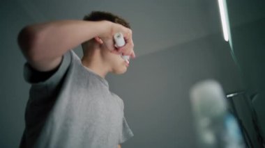 Beyaz çocuk banyoda aynaya bakar, dişlerini fırçalar. Ergen lavabonun önünde duruyor ve yatmadan önce elektrikli diş fırçası kullanıyor. Akşam banyosu rutini. Yaşam tarzı konsepti. Düşük açılı çekim.