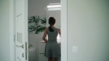 Genç bir kız banyoya gelir, yüzünü soğuk suyla yıkar ve aynaya bakar. Genç lavabonun önünde duruyor ve dişlerini fırçalamak için elektrikli diş fırçası kullanıyor. Kapının arkasından arka plan.