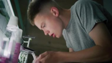 Beyaz çocuk lavaboda duruyor, yüzünü soğuk suyla yıkıyor ve havluyla ellerini siliyor. Genç adam aynadaki yansımasına bakıyor ve sabah banyolarını rutin hale getiriyor. Düşük açı görünümü.