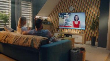Beyaz erkek ve kadın TV 'de haber yayını izliyor, Birleşik Devletler başkanlık seçimlerini tartışıyor ve evdeki kanepede çay içiyor. TV şovları, kadın haber spikeriyle son dakika gelişmelerini sunuyor.