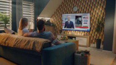 Beyaz çift, ABD başkanlık seçimlerini televizyondaki büyük ekranda izliyor, evdeki kanepede oturup çay içiyor ve konuşuyor. Erkek haber spikerinin TV ekranında yayınladığı son dakika haberi.