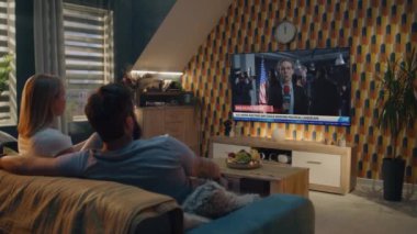 Yetişkin çiftler, evdeki kanepede oturan büyük TV ekranında ABD Seçim Günü ile ilgili haber bülteni izleyip sohbet ediyorlar. TV ekranında beyaz kadın muhabirle ilgili son dakika haberleri yayınlanıyor..