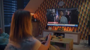 Beyaz kadın, kocasıyla koltukta oturan uzaktan kumandayla ABD başkanlık seçimleriyle ilgili son dakika haberleriyle TV kanallarını değiştirdi. TV ekranında televizyon kanalları ve siyasi programlar gösteriliyor.