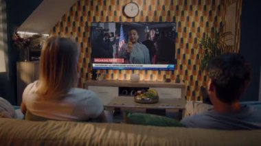 Yetişkin bir çift, Birleşik Devletler seçimleriyle ilgili haber bültenini kanepede oturan büyük TV ekranından izliyor. TV şovu, Afrikalı Amerikalı kadın muhabirin son dakika haberlerini gösteriyor. Arka plan.