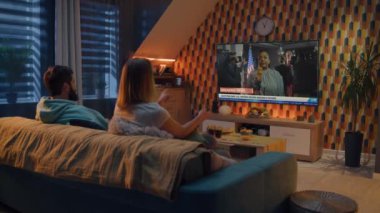 Kafkasyalı çift evlerindeki koltukta oturup TV ekranında Amerika Birleşik Devletleri başkanlık seçimleriyle ilgili haber yayınlarını izliyor. Yetişkin bir kadın uzaktan kumanda kullanarak televizyon kanallarını politik programlarla değiştiriyor..