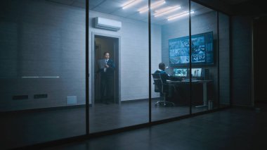 Afrikalı Amerikalı CCTV operatörü bilgisayarla çalışıyor. Ofisteki güvenlik kameralarını izliyor. Birden fazla monitör ve dijital ekrandaki güvenlik kamerası görüntüleri. Bayan çalışan gelir ve iş arkadaşıyla konuşur..