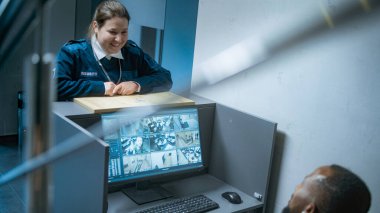 Afrika kökenli Amerikalı erkek güvenlik çalışanı iş yerinde güvenlik kamerası görüntülerini gösteren bir bilgisayar monitörüyle oturuyor, cep telefonu kullanıyor. Kadın gardiyan iş arkadaşıyla konuşur, anahtarları verir. CCTV sistemi.