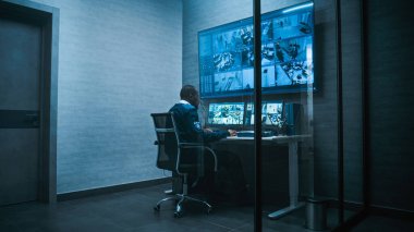 Afrikalı Amerikalı güvenlik görevlisi ofisindeki güvenlik kameralarını bilgisayardan izliyor. Çoklu monitörler ve büyük dijital ekran güvenlik kamerası görüntülerini gösteriyor.