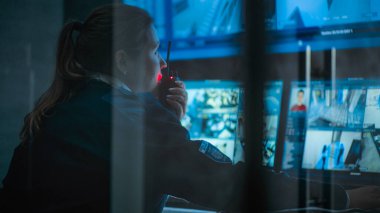 Güvenlik kameralarıyla izleme merkezinde çalışan kadın CCTV operatörünü kapatın, telsizle konuşun. Çoklu monitörler ve güvenlik kamerası görüntülerini gösteren büyük dijital ekran..