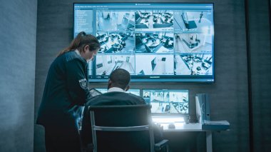 Kadın güvenlik görevlisi Afrikalı Amerikalı iş arkadaşıyla konuşuyor, tablet kullanıyor. Çok ırklı gözetleme operatörleri bilgisayar monitörlerindeki güvenlik kameralarını ve CCTV görüntüleriyle büyük dijital ekranı kontrol ediyorlar..