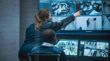 Kadın güvenlik görevlisi Afrikalı Amerikalı meslektaşıyla konuşuyor, tablet kullanıyor. Çok ırklı CCTV operatörleri güvenlik kameralarını bilgisayar monitörlerinde ve güvenlik kamerası görüntüleriyle kontrol ediyorlar..