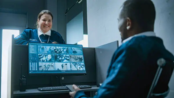 stock image African American male security worker sits at workplace with computer monitor showing surveillance cameras video footage, uses mobile phone. Female guard talks to colleague, gives keys. CCTV system.