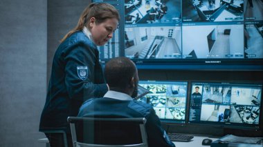 Kadın güvenlik görevlisi Afrikalı Amerikalı meslektaşıyla konuşuyor, tablet kullanıyor. Çok ırklı CCTV operatörleri güvenlik kameralarını bilgisayar monitörlerinde ve güvenlik kamerası görüntüleriyle kontrol ediyorlar..