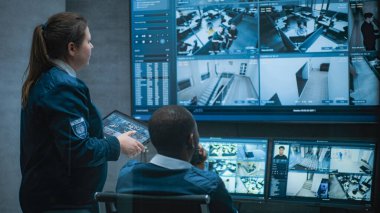 Kadın güvenlik görevlisi Afrikalı Amerikalı meslektaşıyla konuşuyor, tablet kullanıyor. Çok ırklı CCTV operatörleri güvenlik kameralarını bilgisayar monitörlerinde ve güvenlik kamerası görüntüleriyle kontrol ediyorlar..