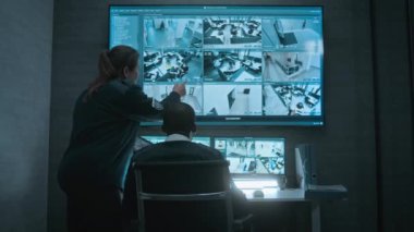 Kadın güvenlik görevlisi Afrikalı Amerikalı iş arkadaşıyla konuşuyor, tablet kullanıyor. Çok ırklı gözetleme operatörleri bilgisayar monitörlerindeki güvenlik kameralarını ve CCTV görüntüleriyle büyük dijital ekranı kontrol ediyorlar..