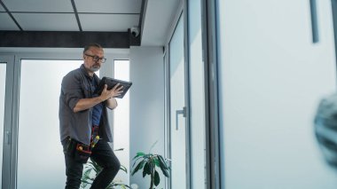 Yetişkin bir erkek teknisyen merdivende durur, güvenlik kameralarını profesyonel yazılımlı tablet bilgisayar kullanarak ofis odasına yerleştirir. İzleme ve izleme. Güvenlik sistemi, video izleme ve mahremiyet.