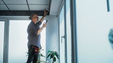 Üniformalı bir erkek, ofis odasındaki güvenlik kamerasını cep telefonunda profesyonel uygulama kullanarak ayarlar. İzleme ve izleme sistemi. Gözetim sistemi, CCTV ve gizlilik kavramı.