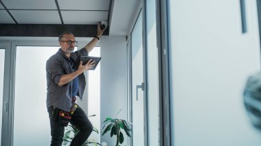 Yetişkin bir erkek teknisyen merdivende durur, güvenlik kameralarını profesyonel yazılımlı tablet bilgisayar kullanarak ofis odasına yerleştirir. İzleme ve izleme. Güvenlik sistemi, video izleme ve mahremiyet.