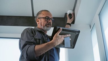 Gözlüklü yetişkin bir erkek, kapalı devre kamerasını tablet bilgisayardaki profesyonel programı kullanarak ofis odasına kuruyor. İzleme ve izleme sistemi. Gözetim sistemi, güvenlik ve gizlilik kavramı.