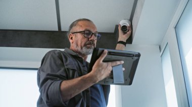 Gözlüklü yetişkin bir erkek, kapalı devre kamerasını tablet bilgisayardaki profesyonel programı kullanarak ofis odasına kuruyor. İzleme ve izleme sistemi. Gözetim sistemi, güvenlik ve gizlilik kavramı.