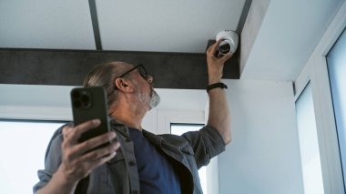 Üniformalı yetişkin erkek teknisyen ofis binasında cep telefonu ve profesyonel uygulama kullanarak güvenlik kamerası kuruyor. İzleme ve izleme sistemi. Gözetim sistemi, güvenlik ve gizlilik kavramı.