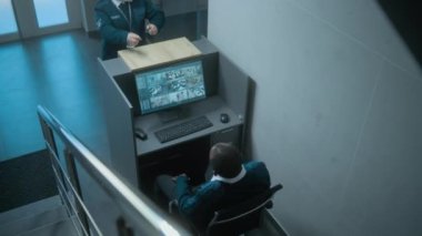 Afrika kökenli Amerikalı erkek güvenlik görevlisi ofisteki iş yerinde oturur, CCTV kameralarından alınan güvenlik görüntüleriyle bilgisayar monitörüne bakar. Kadın işçi gelir ve iş arkadaşıyla konuşur..