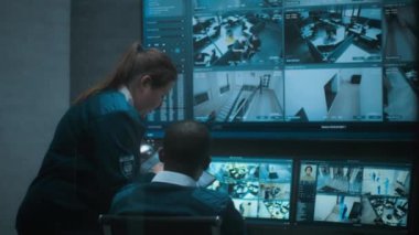 Kadın güvenlik görevlisi Afrikalı Amerikalı meslektaşıyla konuşuyor, tablet kullanıyor. Çok ırklı CCTV operatörleri güvenlik kameralarını bilgisayar monitörlerinde ve güvenlik kamerası görüntüleriyle kontrol ediyorlar..