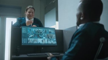 Afrika kökenli Amerikalı erkek güvenlik çalışanı iş yerinde güvenlik kamerası görüntülerini gösteren bir bilgisayar monitörüyle oturuyor, cep telefonu kullanıyor. Kadın gardiyan iş arkadaşıyla konuşur, anahtarları verir. CCTV sistemi.