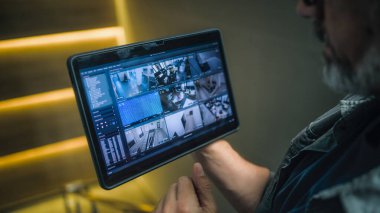 Erkek teknisyen, modern ofisteki güvenlik kameralarını tablet bilgisayar kullanarak izliyor ve kontrol ediyor. Tablet ekranda CCTV kameralarından alınan kayıtlar var. İzleme ve izleme. Güvenlik sistemi ve mahremiyet