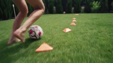 Ev bahçesinde çimlerin üzerinde futbol topunun turuncu koni konilerinin arasından top süren atletik bir çocuğun diz boyu çekimi. Genç futbolcu yarışmaya hazırlanıyor. Hobi ve spor.