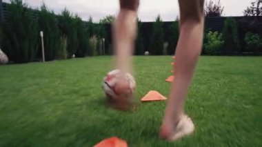 Genç futbolcu, futbol topunu turuncu disk külahlarına sürüyor. Atletik genç çocuk evin bahçesinde çim çimenliğinde antrenman yapıyor, şampiyonluğa hazırlanıyor. Hobi ve spor. Diz hizasında atış.
