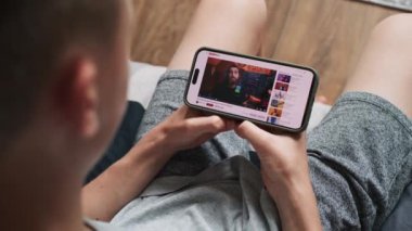 Kafkasyalı çocuk koltukta otururken hisse senedi alım-satımını anlatan eğitici videolar izliyor. Akıllı telefon ekranında video sunucu web arayüzü ile video içeriği gösteriliyor. Kapat..