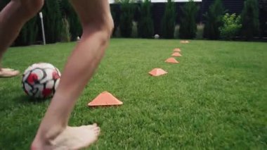 Genç futbolcu turuncu disk konilerinin içinde futbol topu sürüyor. Turnuvaya hazırlanıyor. Atletik genç çocuk evin bahçesinde çimlerin üzerinde antrenman yapıyor. Diz hizasında atış. Yavaş çekim.
