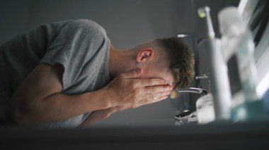 Beyaz çocuk lavaboda duruyor, yüzünü soğuk suyla yıkıyor ve havluyla ellerini siliyor. Genç adam aynadaki yansımasına bakıyor ve sabah banyolarını rutin hale getiriyor. Düşük açı görünümü.