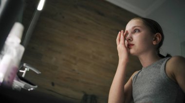 Genç kız banyodaki aynaya bakar ve yüzüne termal su püskürtür. Yumuşak yüzlü güzel kız lavabonun önünde duruyor. Sağlıklı cilt bakımı. Akşam banyosu konsepti.