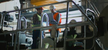 Profesyonel ağır endüstri, güvenlik üniforması, kask ve gözlüklü olgun mühendis dizüstü bilgisayar kullanır ve imalat fabrikasındaki teknisyenlerle konuşur. İki erkek çalışan ekipmanla çalışıyor. Yavaş çekim.