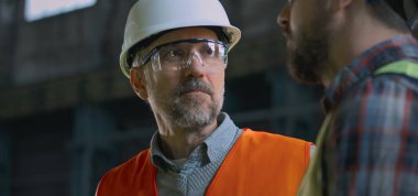 Koruyucu üniformalı, gözlüklü ve miğferli olgun profesyonel mühendis imalat fabrikasında ya da depoda iş arkadaşlarıyla üretim hakkında konuşuyor. Yavaş çekim.