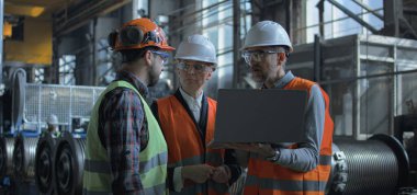 Üretim tesisinde koruyucu üniforma ve kask konusunda üç ağır endüstri uzmanı var. Erkek ve kadın müfettişler dizüstü bilgisayar kullanır ve fabrikadaki işçilerle üretim sürecini tartışırlar. Yavaş çekim.