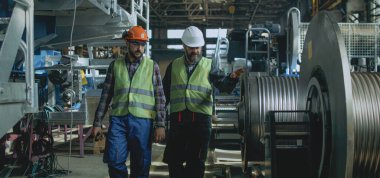 Koruyucu üniformalı ve kasklı iki profesyonel ağır sanayi teknisyeni üretim fabrikasında yürüme süreciyle ilgili tartışıyorlar. Erkek çalışanlar fabrikada çalışıyor. Yavaş çekim.